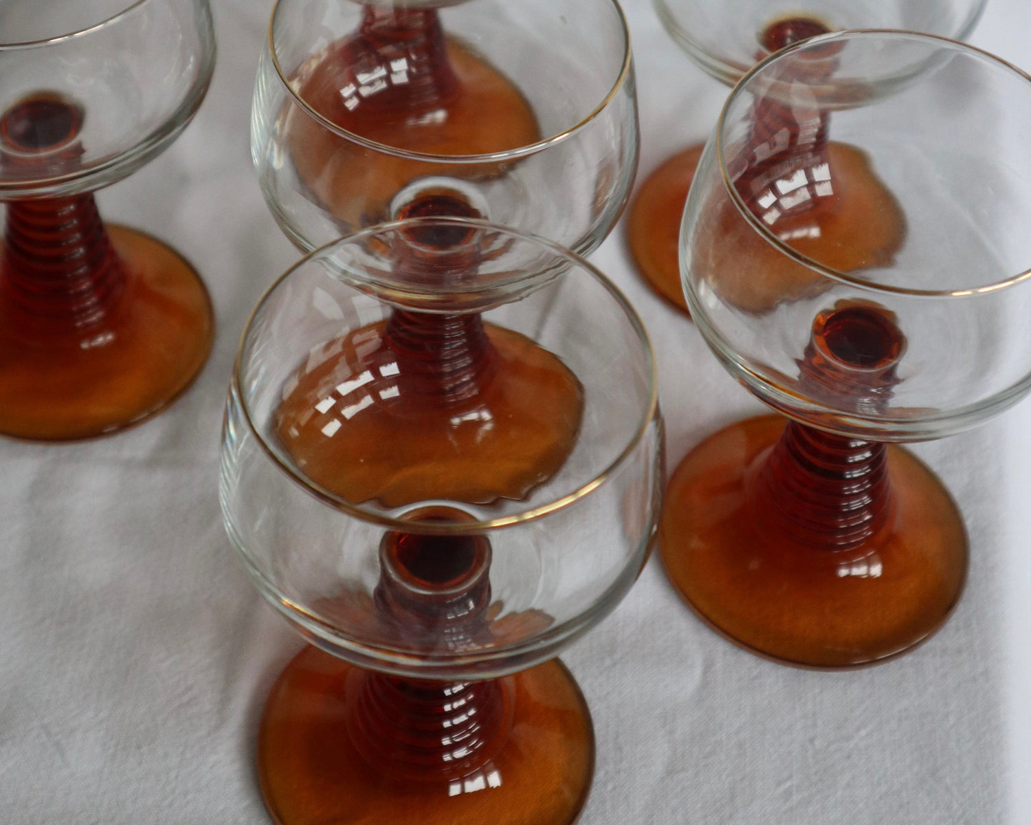 Six clear stemmed glass candle holders with amber-brown ribbed bases, arranged on a white fabric background.
